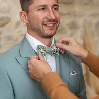 Marié portant un nœud papillon fleuri vert sauge modèle Olivier, fabriqué artisanalement en France, assorti à un costume bleu céladon lors d’un mariage bohème chic et champêtre